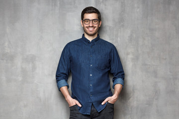Young smiling man wearing trendy glasses and denim shirt, leaning on gray textured wall