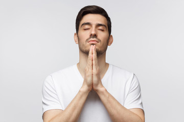 Close-up of young man putting hands together as if he is praying with closed eyes, isolated on gray background