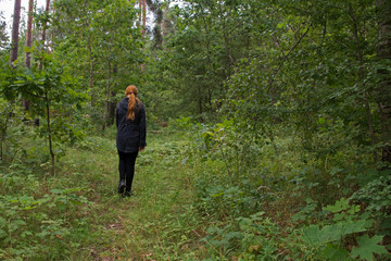Redhaired girl in a black jacket and leggings is on the green grass of the forest among the trees