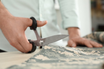 Fototapeta na wymiar Professional tailor cutting camouflage fabric with scissors in workshop, closeup