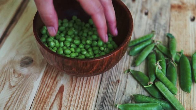 closeup of woman hand depod green peas in brown dish, on wooden table.