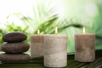 Fototapeta na wymiar Burning candles and spa stones with palm leaf on table against blurred green background