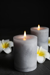 Fototapeta na wymiar Burning candles and plumeria flowers on dark grey table against black background, space for text