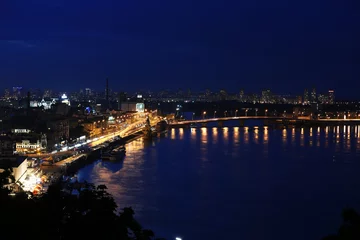 Foto op Plexiglas KYIV, UKRAINE - MAY 21, 2019: Beautiful view of night cityscape with illuminated buildings near river and bridge © New Africa