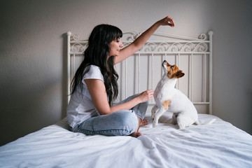 young caucasian woman on bed with her cute small dog playing and giving him treats. Love for animals concept. Lifestyle indoors