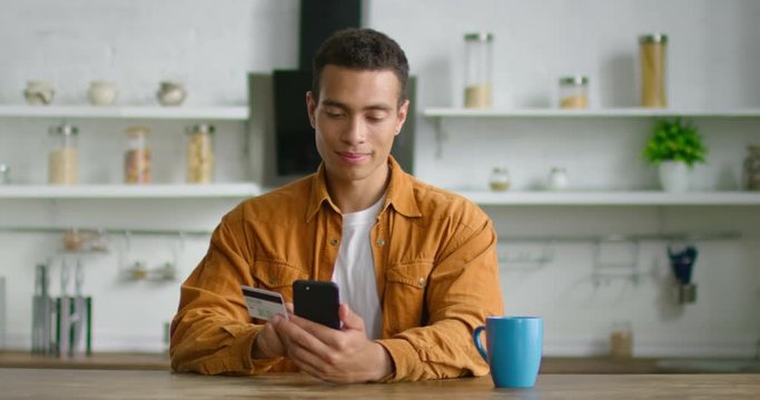 Young Hispanic man is entering credit card number in his smartphone, gadget, doing online shopping, happy and cheerful, sitting at table in kitchen, latin. 4K, shot on RED camera.