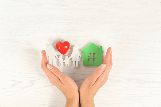 Woman holding hands near red heart and figures of house and family on white wooden background, top view