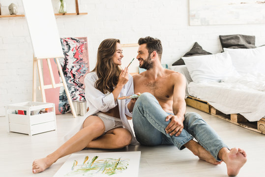 sexy couple sitting on floor and looking at each other while girl trying to paint face of man