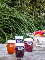 Four jars with homemade jam from blackberries, strawberries and apricots on the table in the garden. Bowl of fresh fruits on the background.