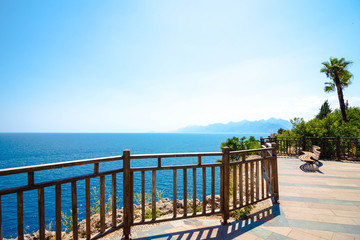 An observation deck overlooking the blue sea. On the site is a bench for rest and tracking with a beautiful view. Palm trees and various bushes grow around.