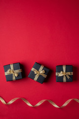 Three black gift box with golden ribbons in line near with twisted ribbon on red paper background, texture, isolated, top view, flat lay, copy space