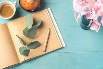 Flat lay, top view of feminine desk. Coffee cup for breakfast, cupcake, empty notebook, branch with eucalyptus leaves and and pink hydrangea. Business planning breakfast concept.