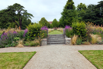 Summer garden with stone path leading to a staircase to wild flower meadow, surrounded by mature trees .