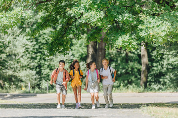 four happy multicultural schoolkids with backpacks running in sunny park