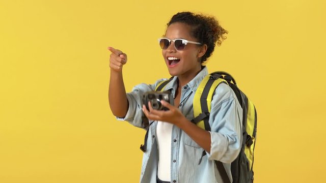 Smiling young African American woman tourist wearing sunglasses and backpack taking photos on camera against yellow backdrop