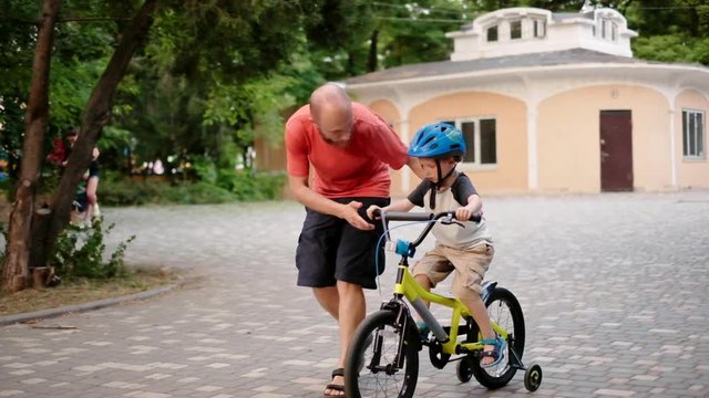 Father teaching son to ride a bike. Man and kid learning to ride bicycle in summer park, having fun together. Parent help child boy riding bicycle outdoors. Active sport family leisure. Slow motion.