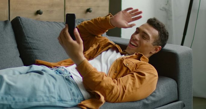 Handsome young Hispanic man is lying on sofa, having video chat on smartphone, gadget, relaxing, in good mood, enjoying weekend in studio apartment. Slow motion, 4K. Shot on RED camera.