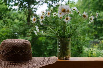 Wicker brown hat and bouquet of daisies in sunny day. Straw cap with chamomiles on rustic wooden terrace. Still life of bunch of white wildflowers with copy space. Summer vacation rural concept