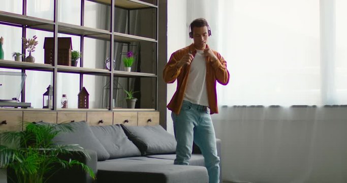Young latin man is dancing in wireless headset in living room, having fun, listening music, enjoying freetime, smiling, being in raised mood, indoor. Hispanic, 4K, shot on RED camera.