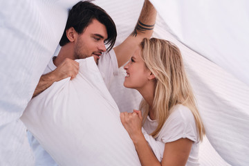 Young attractive brunette man and pretty emotional blond woman thoughtfully looking at each other and fighting for pillow lying together under blanket in bed at cozy home