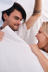 Fototapeta na wymiar Young brunette man and attractive emotional blond woman looking at each other and fighting for pillow lying together under blanket in bed at cozy home