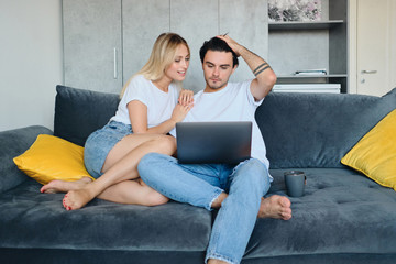 Beautiful smiling blond woman and thoughtful brunette man with cup dreamily using laptop together. Young couple sitting on sofa at modern home