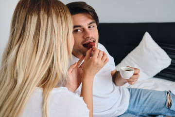 Young brunette man holding cup of coffee in hand dreamily eating strawberry and looking at blond woman feeding him. Couple in white T-shirts having breakfast in bed in hotel