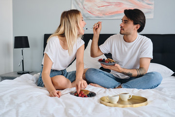 Young brunette man holding plate with berries and pancakes in hand and dreamily feeding pretty blond woman. Beautiful couple in white T-shirts having delicious breakfast in bed in hotel