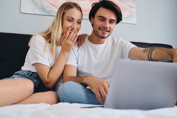 Obraz na płótnie Canvas Beautiful blond woman and smiling brunette man happily watching movie on laptop together. Young beautiful couple in white T-shirts sitting on bed at home