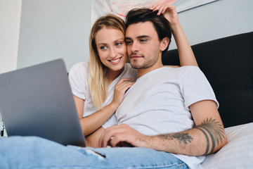 Fototapeta premium Cheerful blond woman happily hugging attractive brunette man while he dreamily working on laptop. Young beautiful couple in white T-shirts sitting on bed together at cozy home