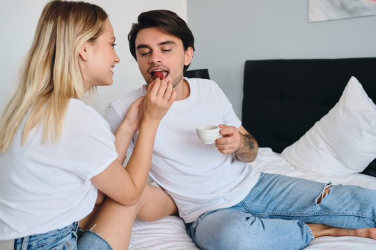 Young Brunette Man Holding Cup Of Coffee In Hand Dreamily Eating Strawberry While Pretty Blond Woman Feeding Him. Beautiful Couple In White T-shirts Having Delicious Breakfast In Bed In Hotel