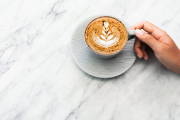 Cup of fresh coffee cappuccino in woman hands on white marble table trendy background. Classic latte art and chocolate on foam. Empty place for text, copy space. Coffee addiction. Top view, flat lay.