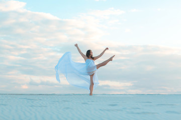 A girl in a fly white dress dances and poses in the sand desert at sunset