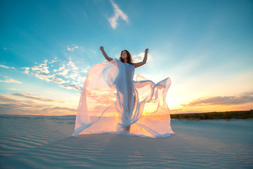 A girl in a fly white dress dances and poses in the sand desert at sunset © Kate