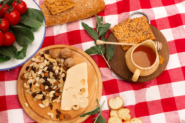 picnic on the street on a checkered blanket spread out food. Honey and nuts on a wooden tray, next to bread, cheese, tomatoes and greens