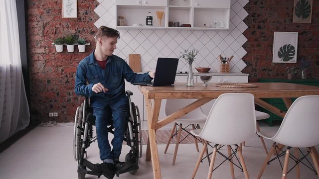 Adult disabled man using his modern computer and spending weekend at cozy bright room in house with interior. Male sitting in special comfort wheelchair making calm glad and smiling face