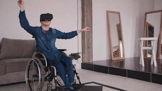 Adult disabled man wearing in virtual reality glasses, sitting in comfort wheel chair and spending free time at home with bright light room in modern interior flat. Male using cyber space interface