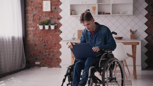 Young or adult disabled man wearing in casual jeans wear, sitting in comfort wheel chair and spending his holiday at cozy house with bright interior room. Male using modern laptop and checking mail