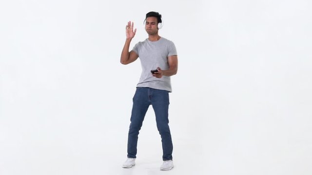 Casual young handsome Indian man holding mobile phone in hand dancing while listening to the music on headphones against white background