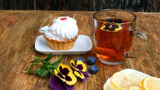 Child hand takes with teaspoon tasty fresh cake on saucer, on wooden table. On table there is also cup with tea, pansy flowers and saucer with lemon.