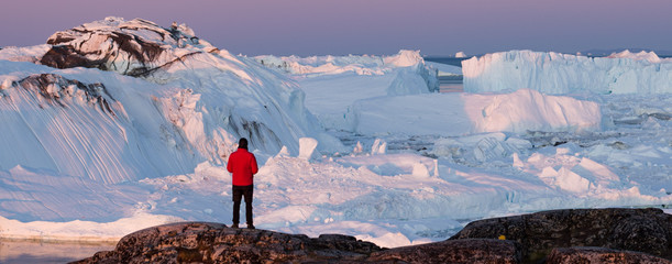 Travel wanderlust adventure in Arctic landscape nature with icebergs - tourist person looking at view of Greenland icefjord - aerial photo. Man by ice and iceberg, Ilulissat Icefjord. - obrazy, fototapety, plakaty