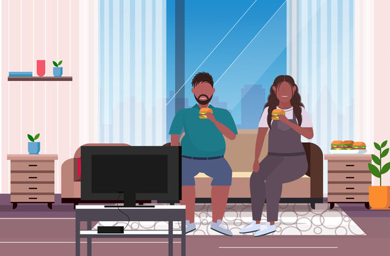 fat obese couple eating hamburger fast food overweight african american man woman watching tv sitting on couch unhealthy nutrition obesity concept living room interior full length flat horizontal