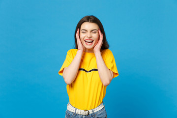 Portrait of laughing young woman in vivid casual clothes keeping eyes closed, putting hands nears face isolated on bright blue wall background in studio. People lifestyle concept. Mock up copy space.