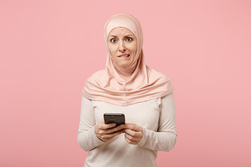 Nervous arabian muslim woman in hijab light clothes posing isolated on pink background. People religious Islam lifestyle concept. Mock up copy space. Using mobile phone typing sms message biting lips.