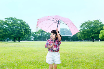 雨の公園で傘を差しテルテル坊主を見つめる幼い女の子。