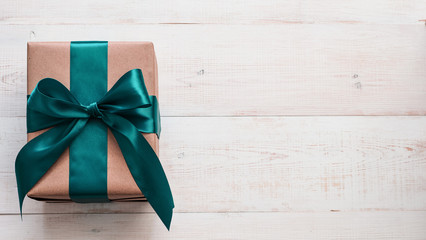 Gift box in craft wrapping paper and green satin ribbon on white wooden table, copy space right....