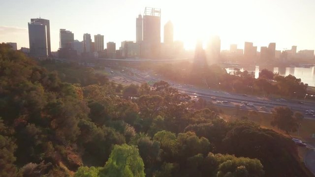 Aerial forward camera motion, flying above the kings park trees and forest towards the sun at sunrise. Revealing busy streets of Perth and morning commute towards Central Business District.