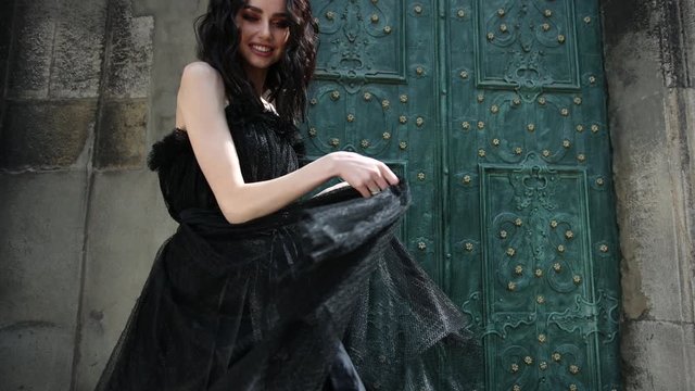 thin beautiful girl, brunette, in a long black dress, with bright make-up, crown on her head, posing for the camera, dancing, on the street near the old building, with high green doors, slow shooting