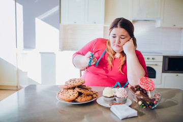 Fat young woman in kitchen sitting and eating sweet food. Upset plus size model look at cookies....