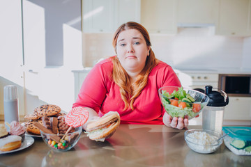 Fat young woman in kitchen sitting and eating food. Confused unhappy plus size model hold burger and bowl salad in hands and look on camera. Alone in kitchen.